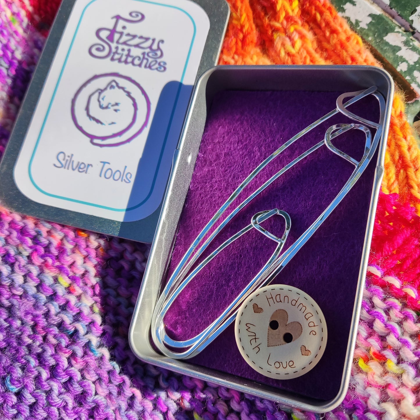 A set of three stitch holders and a button that says "Handmade with love" in a Fizzy Stitches tin, on a background of colourful knitting.