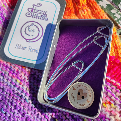 A set of three stitch holders and a button that says "Handmade with love" in a Fizzy Stitches tin, on a background of colourful knitting.