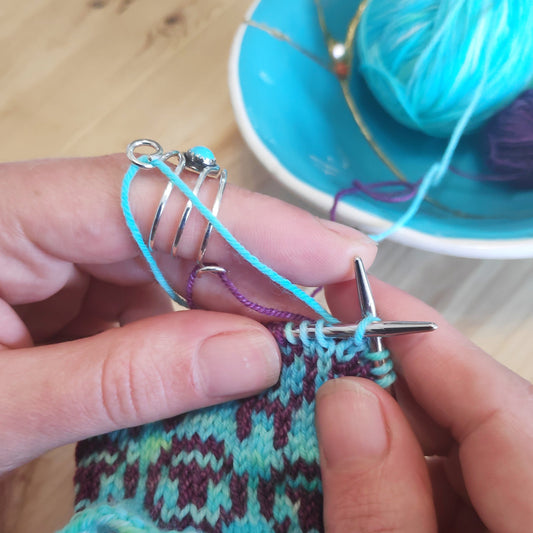 A Sterling silver ring with a turquoise gemstone and two loops, strung with yarn, knitting a colourwork design..
